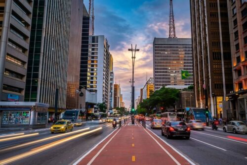 Vista da Ciclovia da Avenida Paulista com rastro de luzes de carros e edifícios ao fundo durante o pôr do sol.