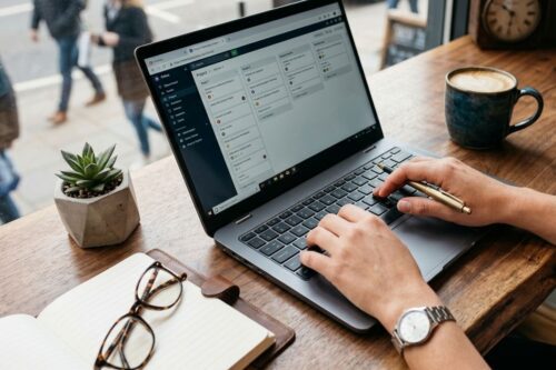 Visão em close das mãos de uma pessoa digitando em um laptop prata sobre uma mesa de madeira rústica.