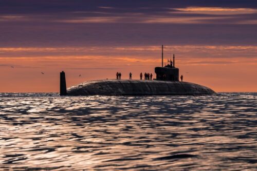 Uma fotografia horizontal com uma perspectiva em nível de olhos mostra um grande submarino russo em primeiro plano, movido a energia nuclear e equipado com mísseis balísticos.