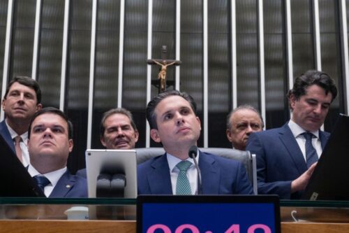 Homem de terno azul e gravata verde fala ao microfone em uma sessão oficial.