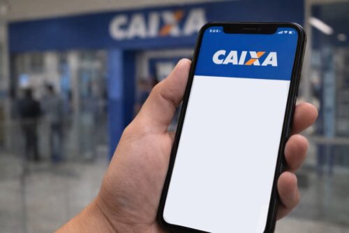 Pessoa segura celular com app da Caixa exibindo erro, em frente a agência bancária desfocada ao fundo.