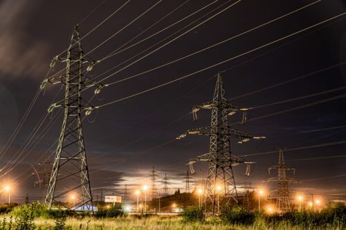 Torres de transmissão de energia à noite, com luzes ao fundo, simbolizando alta nos preços e pressão no setor elétrico.