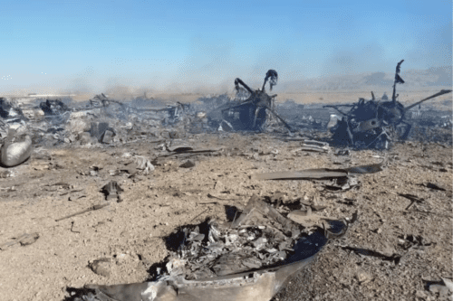 Fotografia em plano aberto de um cenário de destruição em uma área árida e desértica sob um céu azul claro. No solo arenoso e repleto de pedregulhos, veem-se destroços retorcidos e carbonizados de veículos ou aeronaves espalhados por uma grande extensão. Há focos de fumaça cinza subindo dos escombros em vários pontos ao fundo, sugerindo um impacto ou ataque recente. Ao longe, avistam-se formações montanhosas baixas em tons de bege. A iluminação é de luz solar direta e intensa.