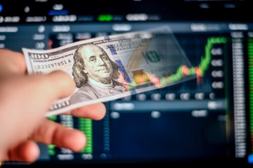 Investidor segurando uma nota de cem dólares, analisando gráfico financeiro com alta, ações e sucesso no mercado de ações.