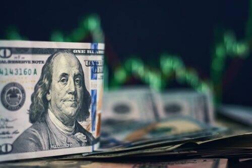 Nota de dólar com gráfico ao fundo ilustra queda da moeda, que fechou abaixo de R$ 5 com cenário externo favorável.