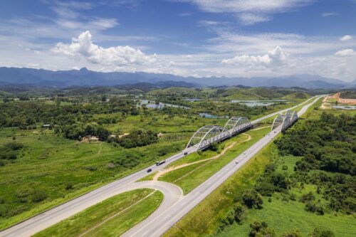 Rodovia com ponte em área verde, cercada por montanhas e rios, sob céu parcialmente nublado, com poucos veículos em circulação.