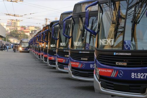 Uma fileira de ônibus urbanos azuis e prateados está estacionada em um terreno pavimentado, com prédios e linhas de energia visíveis ao fundo, durante o Feriado de Tiradentes em São Paulo.
