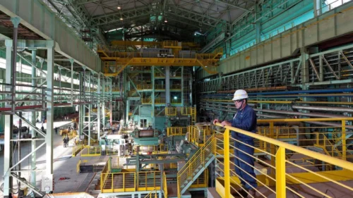 Interior de usina siderúrgica da Usiminas (USIM5), com operador e equipamentos industriais em operação.