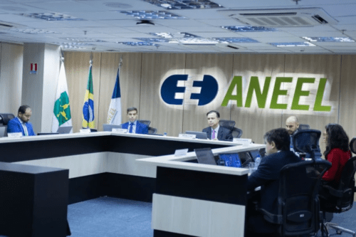 Uma sala de reuniões formal onde ocorre uma sessão ou painel de discussão. Ao fundo, uma parede de madeira clara exibe o logotipo iluminado da ANEEL (Agência Nacional de Energia Elétrica), com as letras "EE" estilizadas em azul e a palavra "ANEEL" em verde. À esquerda, há três bandeiras oficiais em pedestais, incluindo a bandeira do Brasil. Várias pessoas em trajes formais estão sentadas ao redor de uma grande mesa em formato de "U" de cor preta e branca. Alguns participantes, de terno, observam documentos ou telas de notebooks, enquanto outros estão posicionados em primeiro plano, de costas ou de perfil, acompanhando a reunião. O ambiente é bem iluminado por luminárias embutidas no teto comercial.