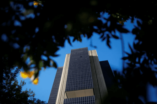 Uma fotografia tirada de baixo para cima mostra a fachada do edifício do Banco Central do Brasil, em Brasília, emoldurada pela silhueta escura de folhas e galhos de árvores em primeiro plano. O prédio imponente de arquitetura brutalista tem colunas verticais de concreto claro intercaladas por grandes janelas de vidro escuro. Ao fundo, o céu está limpo e com um tom azul intenso. Algumas pequenas flores amarelas aparecem desfocadas entre as folhas no canto esquerdo.