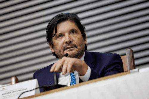 Fotografia em plano médio do deputado André do Prado. Ele é um homem de pele clara, cabelos escuros e levemente grisalhos na barba curta, e veste terno azul-escuro com gravata azul clara texturizada. Ele está sentado atrás de uma bancada branca, falando ao microfone com a mão direita em primeiro plano. Ao fundo, uma parede com detalhes em linhas horizontais cinzas.