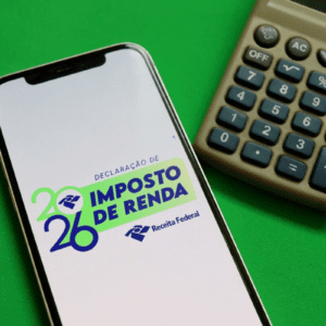 Uma imagem vista de cima sobre um fundo verde sólido mostra um smartphone e uma calculadora analógica marrom posicionada à direita. A tela do celular está acesa e exibe o logotipo oficial da Receita Federal do Brasil para a declaração do Imposto de Renda de 2026. O texto na tela diz "DECLARAÇÃO DE IMPOSTO DE RENDA" em letras maiúsculas nas cores azul e verde, com o ano "2026" estilizado à esquerda e a assinatura da "Receita Federal" logo abaixo.