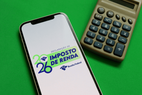 Uma imagem vista de cima sobre um fundo verde sólido mostra um smartphone e uma calculadora analógica marrom posicionada à direita. A tela do celular está acesa e exibe o logotipo oficial da Receita Federal do Brasil para a declaração do Imposto de Renda de 2026. O texto na tela diz "DECLARAÇÃO DE IMPOSTO DE RENDA" em letras maiúsculas nas cores azul e verde, com o ano "2026" estilizado à esquerda e a assinatura da "Receita Federal" logo abaixo.