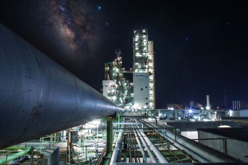 Vista noturna de uma unidade industrial da Braskem. A imagem destaca uma rede complexa de tubulações em primeiro plano que conduz o olhar para torres altas e iluminadas ao fundo.