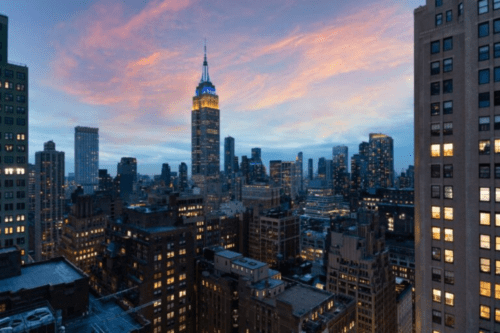 Vista panorâmica do horizonte de Nova York ao entardecer, destacando o Empire State Building iluminado com as cores azul e amarelo contra um céu com nuvens em tons de rosa e laranja. Diversos arranha-céus com janelas acesas preenchem a paisagem urbana sob a luz suave do crepúsculo.