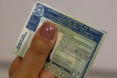 Uma mão segura uma Carteira Nacional de Habilitação (CNH) brasileira aberta.