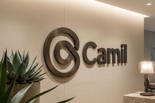 Logotipo da empresa Camil em letras metálicas de bronze fixado em uma parede bege texturizada.