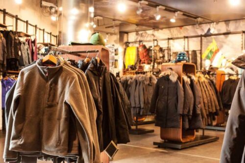 Interior de uma loja de vestuário focada em moda de inverno e atividades ao ar livre. Vários casacos pesados, jaquetas térmicas e parkas em tons de marrom, cinza e preto estão expostos em araras e suportes de madeira.