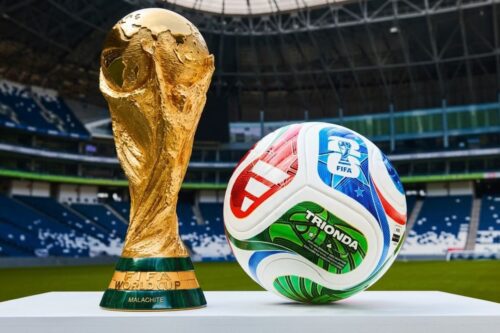 Uma fotografia em alta resolução e ângulo baixo mostrando o Troféu da Copa do Mundo da FIFA em ouro brilhante posicionado à esquerda de uma bola de futebol colorida.