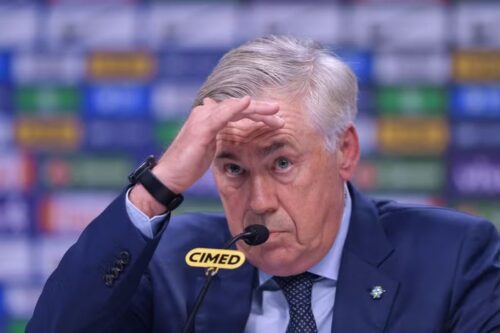 O técnico de futebol Carlo Ancelotti durante uma coletiva de imprensa. Ele veste um terno azul escuro e camisa clara, com a mão direita na testa em um gesto de reflexão ou cansaço.