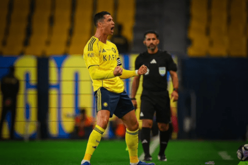 Cristiano Ronaldo, jogador de futebol profissional, em pé no campo durante uma partida, comemorando intensamente com os punhos cerrados e a boca aberta em um grito de comemoração. Ele veste o uniforme amarelo e azul do Al-Nassr FC, composto por uma camisa amarela de mangas compridas, calções azuis e meias amarelas. Ao fundo, um árbitro com uniforme preto observa a cena, e as arquibancadas do estádio estão parcialmente visíveis nas cores amarelo e azul desfocadas.