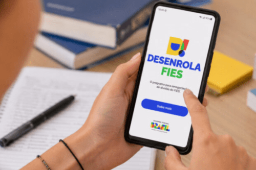 Foto em plano detalhado mostrando as mãos de uma pessoa segurando um smartphone na vertical sobre uma mesa de estudos. Na tela do celular, exibe-se a página do programa "Desenrola Fies", com uma identidade visual colorida em tons de azul, amarelo, verde e vermelho. Abaixo do logotipo, lê-se o texto informativo "O programa para renegociação de dívidas do FIES", seguido por um grande botão azul escrito "Saiba mais" e, no rodapé, a marca do Governo Federal do Brasil. Ao fundo, fora de foco, aparecem um caderno aberto com uma caneta preta ao lado e alguns livros empilhados sobre a mesa de madeira clara.