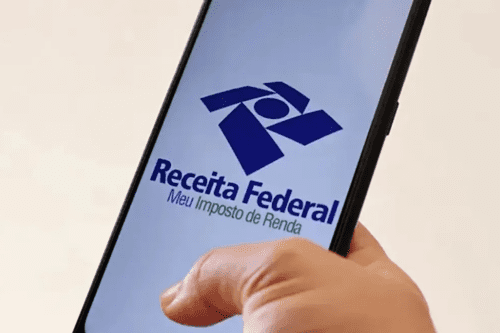 Uma pessoa segura um smartphone inclinado na diagonal contra um fundo claro. Na tela do celular, destaca-se o logotipo azul da Receita Federal do Brasil, composto por formas geométricas estilizadas. Logo abaixo do logo, está escrito em letras azuis "Receita Federal" e, em letras verdes menores, "Meu Imposto de Renda". O polegar da pessoa toca a parte inferior da tela.