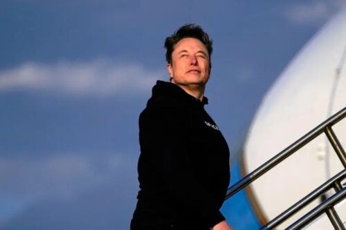 Elon Musk visto de perfil e de baixo para cima, olhando para o horizonte com uma expressão séria.
