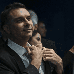 Fotografia em plano médio do político brasileiro Flávio Bolsonaro. Ele veste terno escuro, camisa branca e gravata clara, e aparece ajustando o colarinho com as duas mãos, exibindo uma aliança dourada no dedo anelar esquerdo. Ele olha para o lado com um leve sorriso. O fundo é escuro e traz outras pessoas desfocadas, com destaque para uma mão segurando um smartphone rosa tirando uma foto.