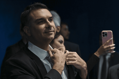 Fotografia em plano médio do político brasileiro Flávio Bolsonaro. Ele veste terno escuro, camisa branca e gravata clara, e aparece ajustando o colarinho com as duas mãos, exibindo uma aliança dourada no dedo anelar esquerdo. Ele olha para o lado com um leve sorriso. O fundo é escuro e traz outras pessoas desfocadas, com destaque para uma mão segurando um smartphone rosa tirando uma foto.