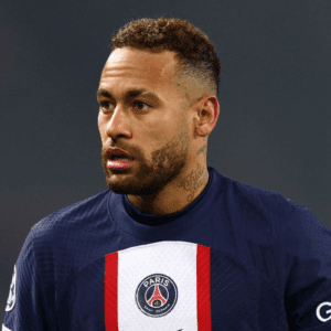 Uma imagem em plano médio centraliza o jogador de futebol Neymar Jr. do peito para cima, olhando ligeiramente para a esquerda com uma expressão séria e concentrada. Ele tem pele parda, barba curta e bem aparada, e cabelo castanho curto, raspado nas laterais em estilo fade (degradê) e texturizado no topo.