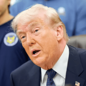 Uma imagem em plano médio foca no rosto de Donald Trump de perfil de três quartos, olhando para a esquerda com a boca aberta como se estivesse falando. Ele tem cabelos loiros claros penteados para trás e pele bronzeada. Ele usa um paletó de terno azul-marinho, camisa social branca e uma gravata azul-escura com bolinhas vermelhas e brancas. Um pequeno broche da bandeira dos Estados Unidos está preso na lapela esquerda de seu paletó. À esquerda, em segundo plano, parte do tronco de um jovem é visível, vestindo uma camiseta azul-escura com um logotipo circular amarelo e azul no peito. O fundo está ligeiramente desfocado, mostrando tons de azul e um vislumbre de um objeto vermelho à direita. A iluminação é clara e direta, destacando as feições de Trump.