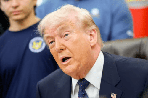 Uma imagem em plano médio foca no rosto de Donald Trump de perfil de três quartos, olhando para a esquerda com a boca aberta como se estivesse falando. Ele tem cabelos loiros claros penteados para trás e pele bronzeada. Ele usa um paletó de terno azul-marinho, camisa social branca e uma gravata azul-escura com bolinhas vermelhas e brancas. Um pequeno broche da bandeira dos Estados Unidos está preso na lapela esquerda de seu paletó. À esquerda, em segundo plano, parte do tronco de um jovem é visível, vestindo uma camiseta azul-escura com um logotipo circular amarelo e azul no peito. O fundo está ligeiramente desfocado, mostrando tons de azul e um vislumbre de um objeto vermelho à direita. A iluminação é clara e direta, destacando as feições de Trump.
