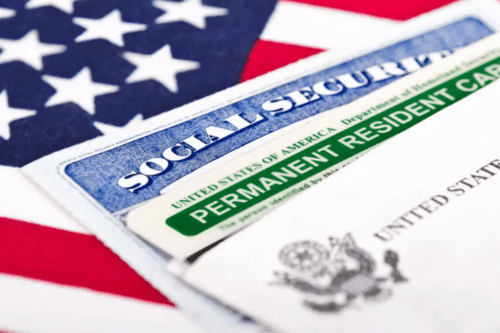 Uma imagem em ângulo fechado mostra um cartão de Residência Permanente dos Estados Unidos (Green Card) e um cartão do Social Security sobrepostos em cima de uma bandeira americana. Os documentos estão ligeiramente desfocados nas extremidades, com foco principal no texto "Permanent Resident Card".