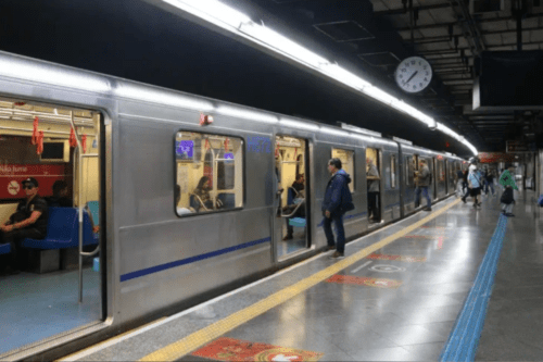 Fotografia do interior de uma estação de metrô em São Paulo. Um trem de metal prateado está parado na plataforma com as portas abertas. Algumas pessoas são vistas dentro dos vagões e outras poucas caminham pela plataforma, que possui piso cinza brilhante, faixa amarela de segurança e sinalização tátil azul. No alto, à direita, um relógio analógico marca aproximadamente 10h10. A iluminação é feita por longas lâmpadas fluorescentes no teto.