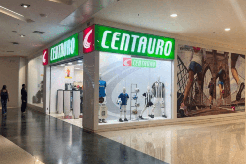 Fachada de uma loja de artigos esportivos Centauro localizada no interior de um shopping center. O letreiro luminoso exibe o nome "CENTAURO" em letras brancas sobre um fundo verde vibrante, acompanhado pelo logotipo vermelho da marca. Na vitrine principal, manequins adultos e infantis vestem uniformes de futebol, cercados por bolas esportivas. À direita, um painel publicitário de grandes dimensões mostra pessoas correndo em uma ponte. O piso do corredor do shopping é polido e reflete a iluminação interna, com algumas pessoas caminhando ao fundo.