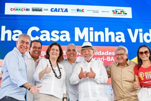 Fotografia de um grupo de sete pessoas sorridentes em um evento oficial. No centro, o presidente Lula usa um chapéu claro e faz um gesto de "positivo" com as duas mãos. Ao fundo, um painel azul com os logotipos do Governo Federal, Caixa, Novo PAC e a frase parcial "Minha Casa Minha Vida".