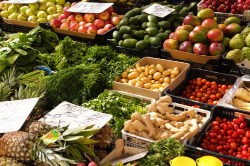Vista superior de uma banca de feira ou hortifrúti repleta de alimentos frescos, ilustrando a variação de preços de itens essenciais do cotidiano. Em primeiro plano, destacam-se abacaxis, gengibre, ervas aromáticas e caixas com tomates-cereja e tomates maduros. Ao centro, há caixas com batatas e maços de temperos verdes como coentro e salsa. Ao fundo, estão organizadas fileiras de frutas como maçãs verdes e vermelhas, abacates, mangas e uvas, algumas delas acompanhadas de pequenas placas de preços manuscritas.