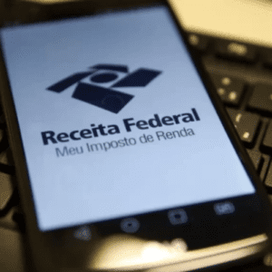 Smartphone apoiado sobre o teclado de um computador, exibindo na tela o logotipo oficial da Receita Federal do Brasil com o texto "Meu Imposto de Renda"
