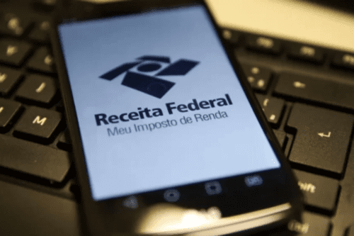 Smartphone apoiado sobre o teclado de um computador, exibindo na tela o logotipo oficial da Receita Federal do Brasil com o texto "Meu Imposto de Renda"
