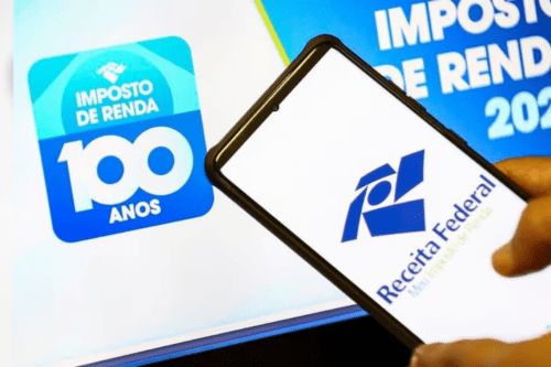 Uma pessoa segura um smartphone em primeiro plano, posicionado diagonalmente. Na tela branca do celular, destaca-se o logotipo azul da Receita Federal do Brasil com o texto "Receita Federal" e, logo abaixo em letras menores, "Meu Imposto de Renda". Ao fundo, a tela de um computador exibe uma imagem azul e branca com um selo comemorativo onde se lê "Imposto de Renda - 100 anos" à esquerda, e parte do texto "Imposto de Renda 202..." à direita.