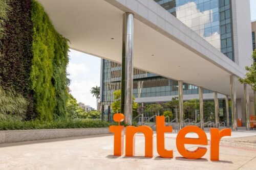 Área externa da sede do Banco Inter. Em primeiro plano, o logotipo "inter" em letras tridimensionais laranjas gigantes está posicionado no chão de uma praça cimentada.