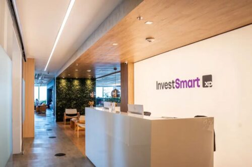 Recepção moderna do escritório da InvestSmart XP. Em destaque, à direita, há um balcão branco minimalista à frente de uma parede branca com o logotipo "InvestSmart XP" em roxo e preto.