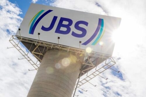 Um grande painel publicitário com o logotipo da empresa JBS no topo de uma torre cilíndrica de concreto, visto de baixo para cima.