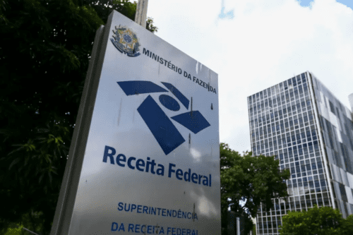 A imagem mostra um totem vertical metálico e escovado da Receita Federal, órgão vinculado ao Ministério da Fazenda do Brasil. O totem está posicionado em primeiro plano, ligeiramente inclinado para a esquerda. Na parte superior, há o Brasão de Armas do Brasil seguido pelo texto "MINISTÉRIO DA FAZENDA". Logo abaixo, destaca-se o logotipo estilizado da Receita Federal em azul escuro e o nome do órgão. Na parte inferior do totem, lê-se "SUPERINTENDÊNCIA DA RECEITA FEDERAL". Ao fundo, do lado esquerdo, observam-se copas de árvores verdes e frondosas. Do lado direito, ergue-se um edifício comercial alto com fachada espelhada e quadriculada, refletindo o céu parcialmente nublado. A foto foi registrada em um ângulo de baixo para cima, sob luz natural diurna.