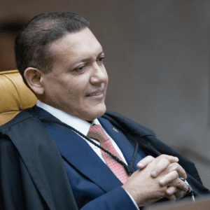 Fotografia em plano médio do Ministro Kassio Nunes Marques sentado no plenário do STF. Ele é um homem de pele parda, cabelos curtos e escuros, e sorri levemente olhando para o lado. Veste uma toga preta sobre um terno azul, camisa branca e gravata coral com padrões discretos. Ele está com as mãos entrelaçadas à frente do corpo e sentado em uma poltrona de couro amarelado com encosto alto. O fundo está desfocado, sugerindo um ambiente formal de escritório ou tribunal.