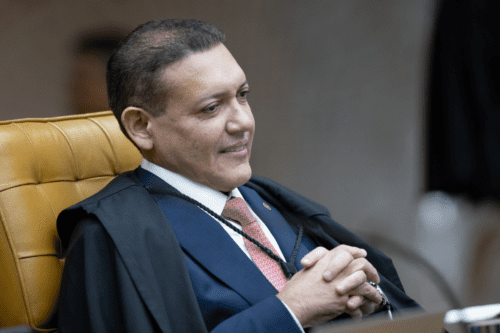 Fotografia em plano médio do Ministro Kassio Nunes Marques sentado no plenário do STF. Ele é um homem de pele parda, cabelos curtos e escuros, e sorri levemente olhando para o lado. Veste uma toga preta sobre um terno azul, camisa branca e gravata coral com padrões discretos. Ele está com as mãos entrelaçadas à frente do corpo e sentado em uma poltrona de couro amarelado com encosto alto. O fundo está desfocado, sugerindo um ambiente formal de escritório ou tribunal.