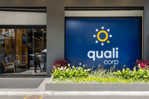 Fachada da entrada de um escritório da Qualicorp. À direita, há um grande painel azul escuro com o logotipo da empresa: um círculo amarelo central cercado por pequenos pontos brancos dispostos em formato de sol, com o nome "quali corp" escrito em letras brancas logo abaixo.