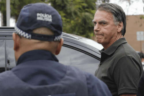 Em primeiro plano, vê-se a parte de trás da cabeça e dos ombros de um policial militar de costas, vestindo uma farda escura e um boné onde se lê "POLÍCIA MILITAR" em letras brancas. À direita, em plano médio e de perfil, o ex-presidente Jair Bolsonaro veste uma camisa polo verde-escura. Ele tem uma expressão séria, com os lábios cerrados, e olha em direção ao policial. Ao fundo, há um veículo escuro e uma vegetação levemente desfocada.