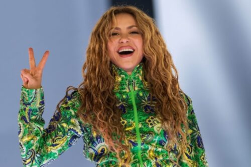 Fotografia em plano médio da cantora Shakira sorrindo e fazendo o sinal de 'v' com os dedos.